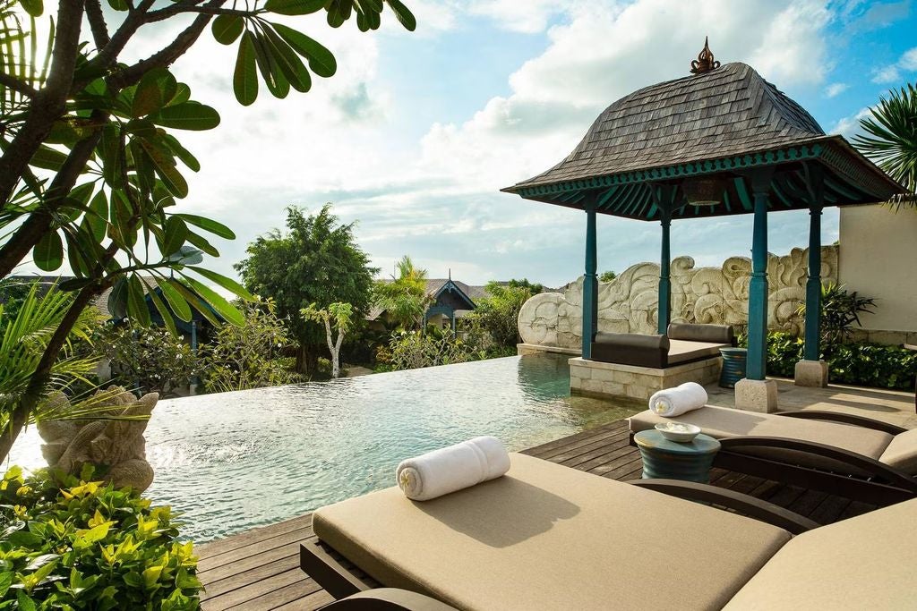 Luxurious villa bedroom with ocean views, private infinity pool, traditional Balinese decor, and floor-to-ceiling windows at sunset