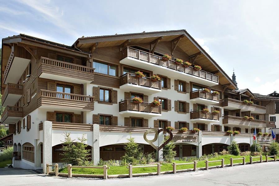 Five-star alpine hotel exterior with stone and wood facade, mountain backdrop, manicured gardens, and elegant balconies in La Clusaz village