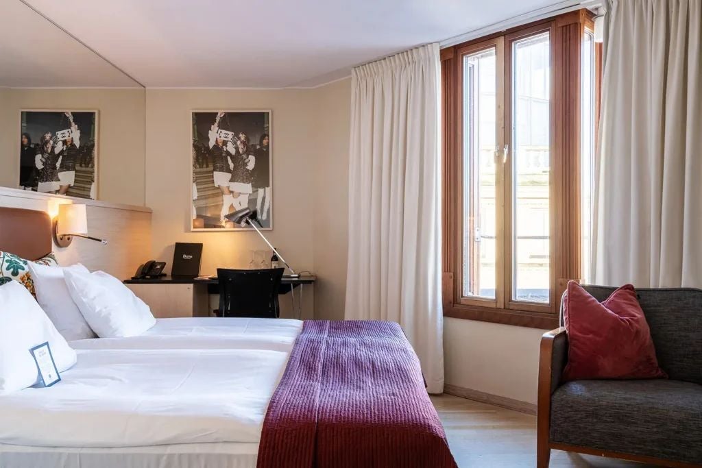 Elegant Swedish hotel room with soft neutral tones, plush king bed, minimalist wooden furniture, and large window overlooking urban Stockholm landscape