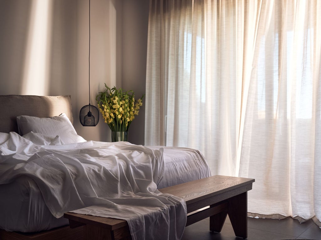 Spacious white-walled Greek villa bedroom with minimalist decor, hardwood floors, blue sea view, and elegant four-poster king bed in soft neutral tones