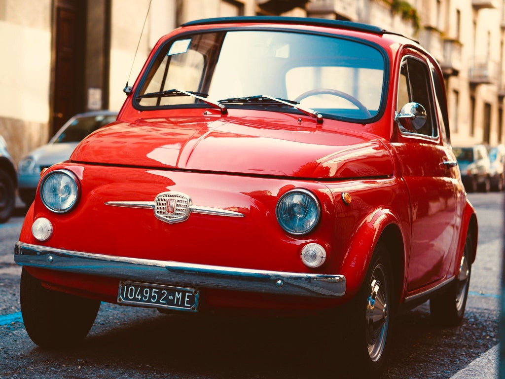 Two vintage Fiat 500 cars in vibrant red and blue parked in front of a rustic Tuscan villa, surrounded by cypress trees and rolling hills