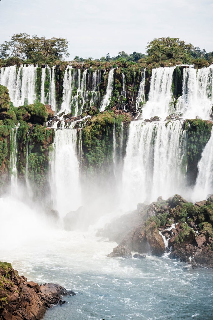 Cascading waterfalls thunder through lush rainforest at Iguazu Falls, mist rising from powerful waters under an overcast sky