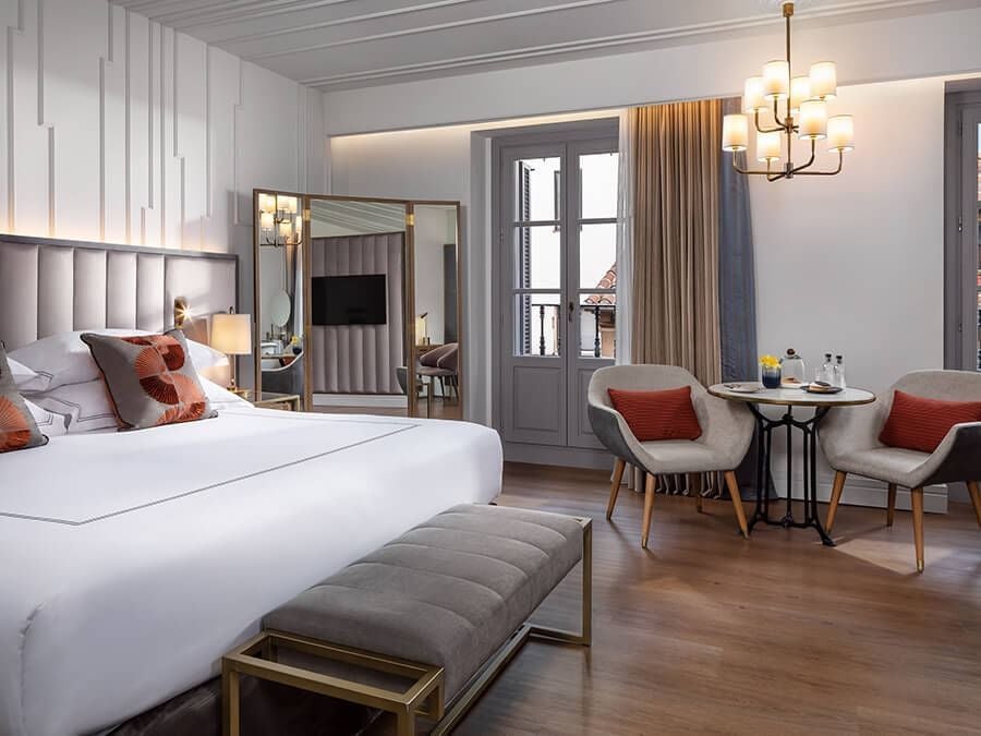 Elegant premium hotel room with plush king bed, vintage-inspired decor, warm wood furnishings, and soft neutral color palette in historic Spanish hotel