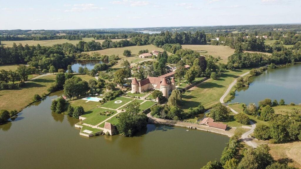 Luxurious French château hotel nestled in lush green countryside, stone architecture with elegant windows, surrounded by serene water and verdant landscape at dusk