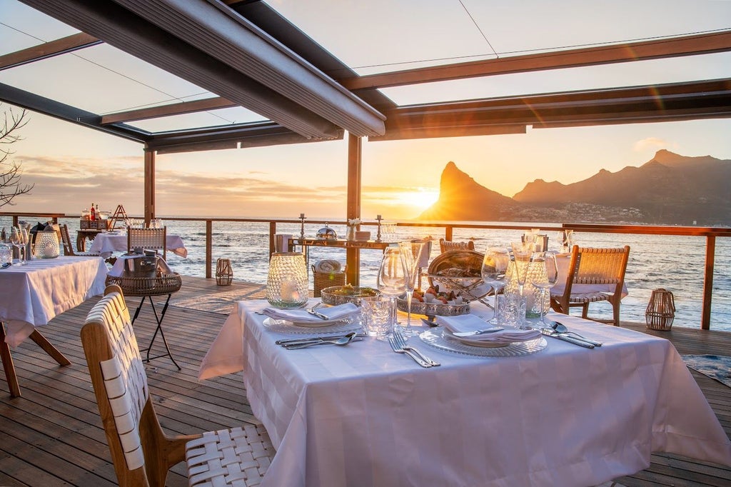 Luxury beachfront lodge with infinity pool overlooking ocean at sunset, nestled beneath mountain peaks along Cape Town's coastline