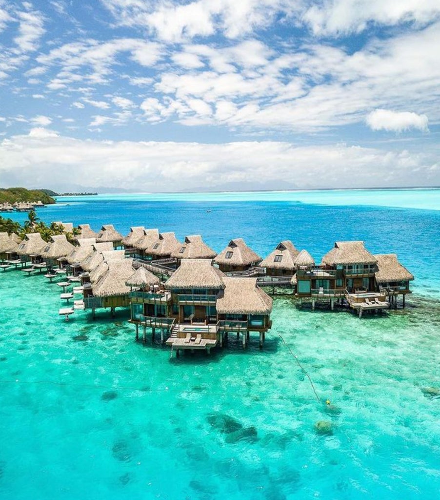 Luxurious overwater bungalows with thatched roofs extend into turquoise Bora Bora lagoon, framed by lush mountains and coral reefs