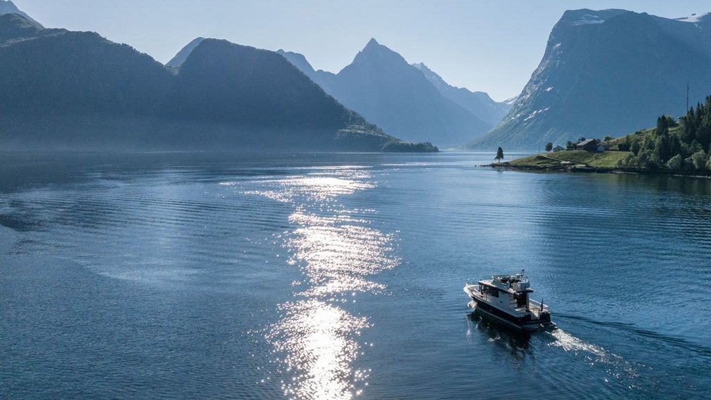 Small luxury yacht cruising through dramatic Norwegian fjord, surrounded by towering cliffs and pristine blue-green waters under clear skies