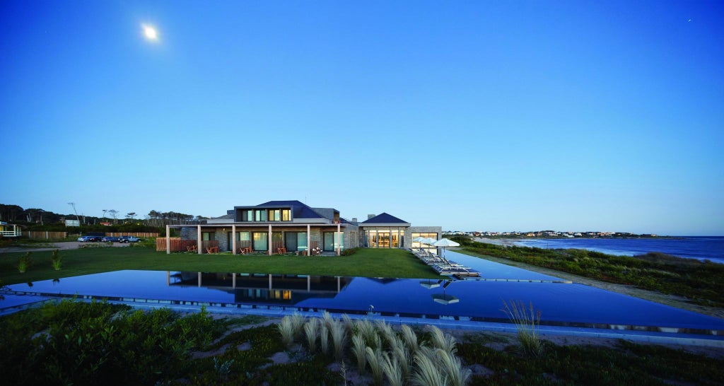 Luxurious beachfront resort with minimalist architecture, blending concrete and glass, overlooking pristine sandy beach and turquoise waters of Uruguay's coastline