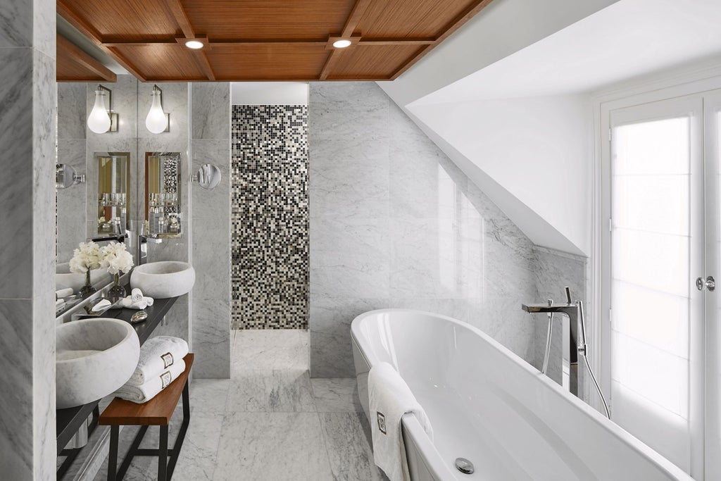 Elegant Parisian suite with plush king bed, designer grey armchair, marble bathroom, and city view through large windows in luxury Hotel Montalembert