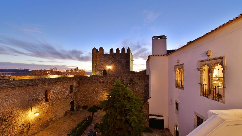 Stone castle hotel with medieval towers and fortified walls perched atop hill overlooking Óbidos village, lit dramatically at dusk