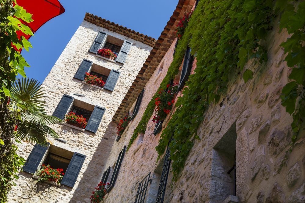 Elegant stone facade of boutique hotel with blue-gray shutters, nestled in charming French village street with cobblestone path and vibrant flowers