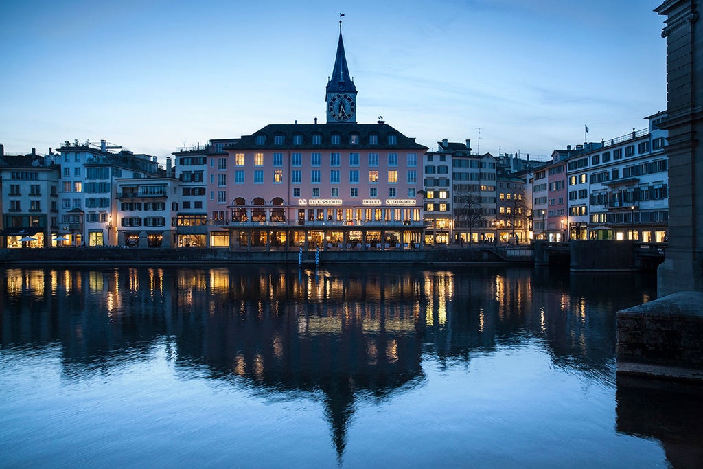 Historic luxury hotel with white facade and red shutters overlooking Limmat River in Zurich, featuring ornate balconies and flags