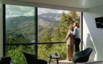 Minimalist wooden suite overlooking dramatic mountain landscape, floor-to-ceiling windows, sleek design, infinity pool merging with rugged Spanish terrain