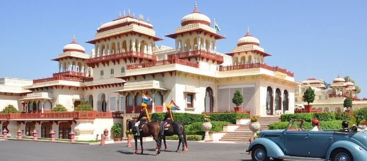 Opulent Rambagh Palace hotel in Jaipur features ornate sandstone archways, manicured gardens, and regal Indian architecture at sunset