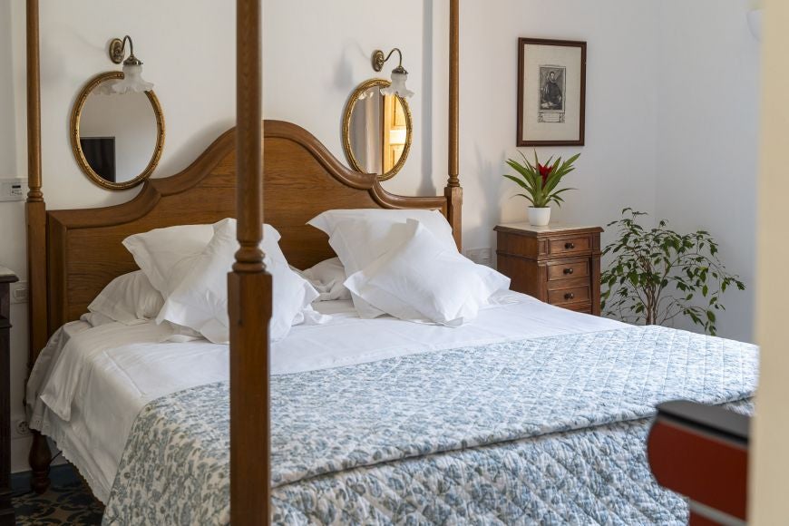 Elegant Spanish luxury bedroom with plush white bedding, soft neutral tones, wooden floors, and panoramic windows overlooking lush Mediterranean landscape scenery