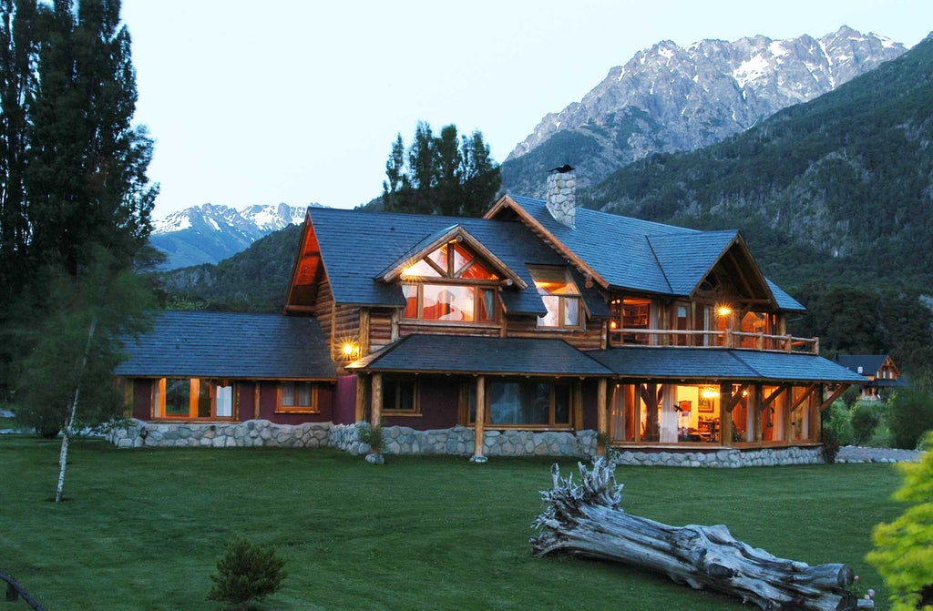 Rustic-luxe lodge with stone and timber facade nestled on Lake Nahuel Huapi, backed by snow-capped Patagonian mountains at sunset