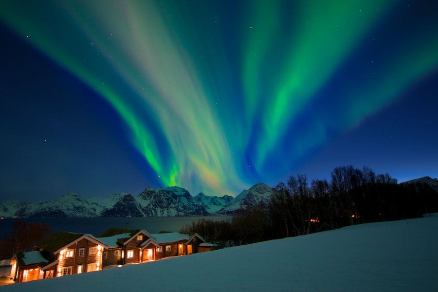 Luxurious wooden lodge nestled in snowy Norwegian mountains, featuring large glass windows reflecting dramatic fjord landscape at sunset