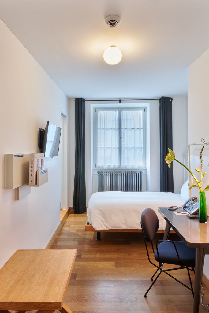 Minimalist Swiss hotel room with modern design, clean white linens, wooden floor, sleek furniture, and large window overlooking urban scenery in Zurich