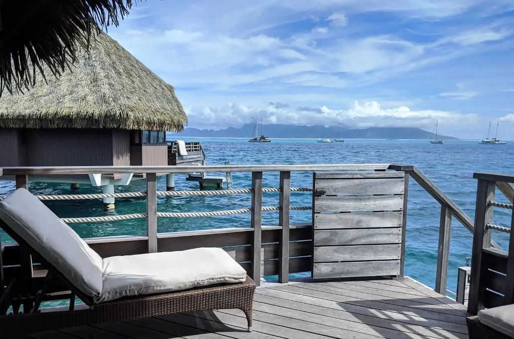 Modern overwater bungalow with private deck, floor-to-ceiling windows overlooking turquoise Tahitian lagoon and Mount Otemanu at sunset