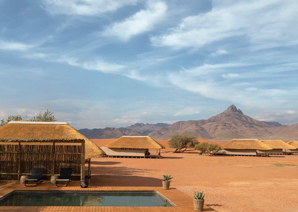 Desert luxury lodge with thatched-roof cottage overlooking vast red sand dunes of NamibRand, golden light illuminating dramatic landscape