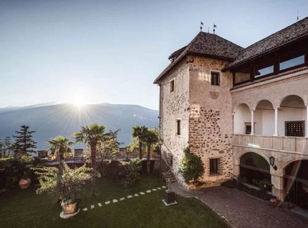 Historic luxury mountain hotel with stone facade and balconies nestled on Italian hillside, overlooking lush valley at sunset