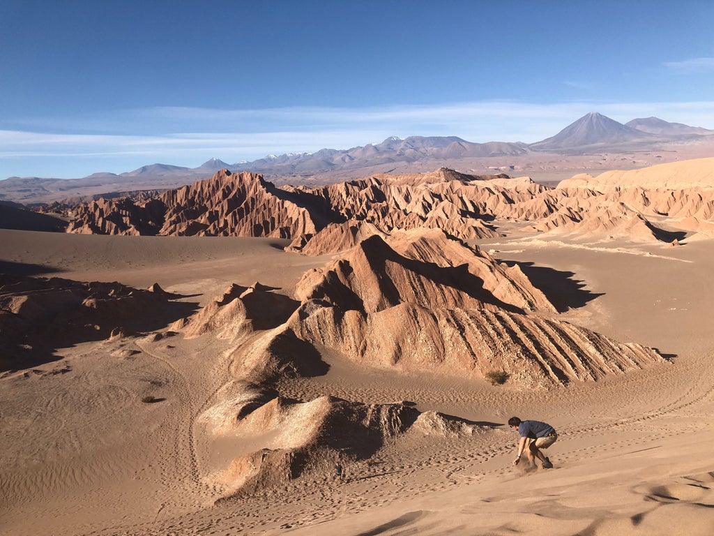 Desert landscape at sunset with rugged mountains silhouetted against orange sky, white luxury tent in foreground amid sand dunes