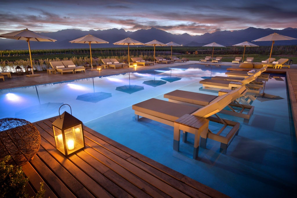 Luxurious stone and glass resort nestled in Argentinian vineyard, with Andes mountains backdrop and infinity pool at sunset