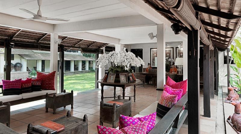 Elegant boutique hotel entrance with wooden gates and stone pathway, framed by lush tropical gardens and traditional Thai architecture