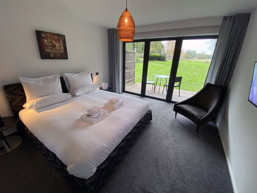 Elegant water meadow-themed hotel room with soft neutral tones, plush bedding, rustic wooden furniture, and tranquil countryside view through large window