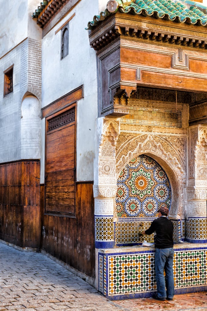 Intricate blue and white ceramic tiles line winding alleyways of a historic Moroccan Medina, with ornate doorways and traditional architecture bathed in golden sunlight.