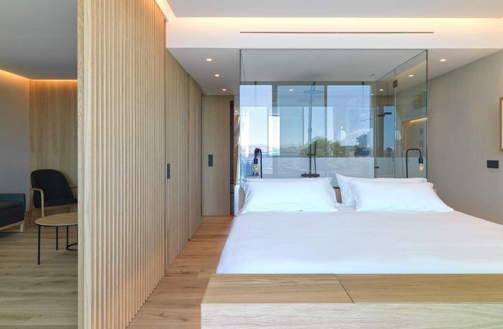 Minimalist white and wooden hotel room with floor-to-ceiling windows, elegant modern furnishings, and sweeping view of Spanish coastal landscape