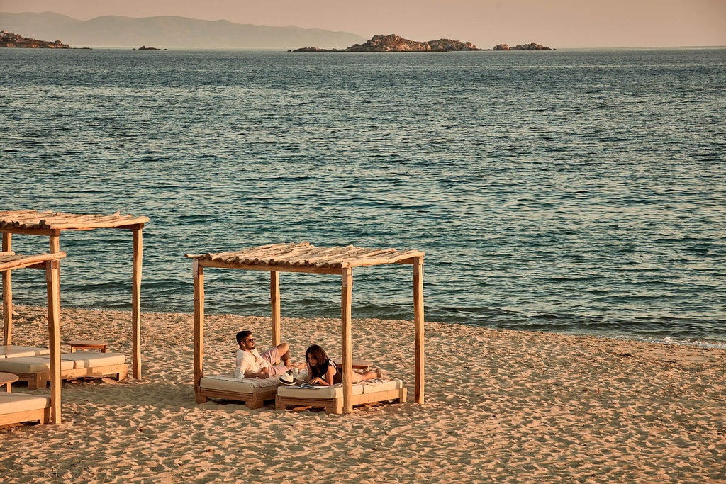 Luxurious beachfront resort with white-washed architecture, azure Aegean waters, and elegant sunbeds overlooking pristine Naxian coastline at sunset