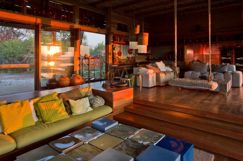 Elevated luxury safari lodge with thatched roof pavilions on wooden platforms, overlooking flooded Okavango Delta grasslands at sunset