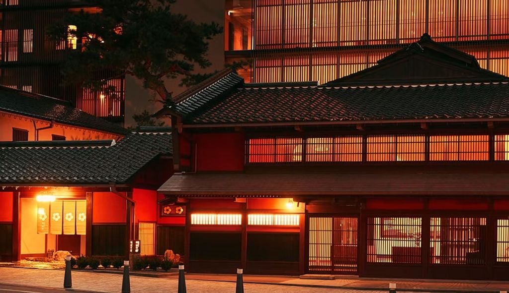 Traditional Japanese ryokan exterior with wooden facade, sliding doors, illuminated lanterns and curved tile roof overlooking garden at dusk