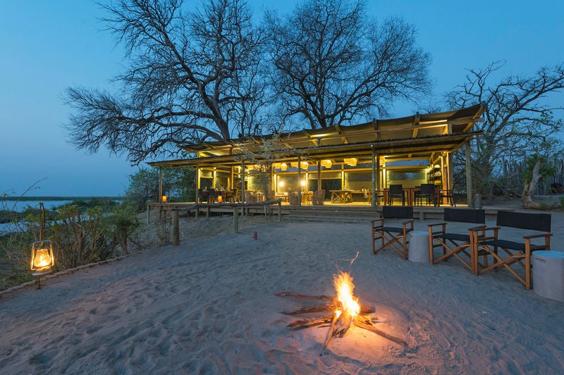 Elevated luxury safari tent with wooden deck overlooking African savanna, palm trees and natural waterhole at golden hour