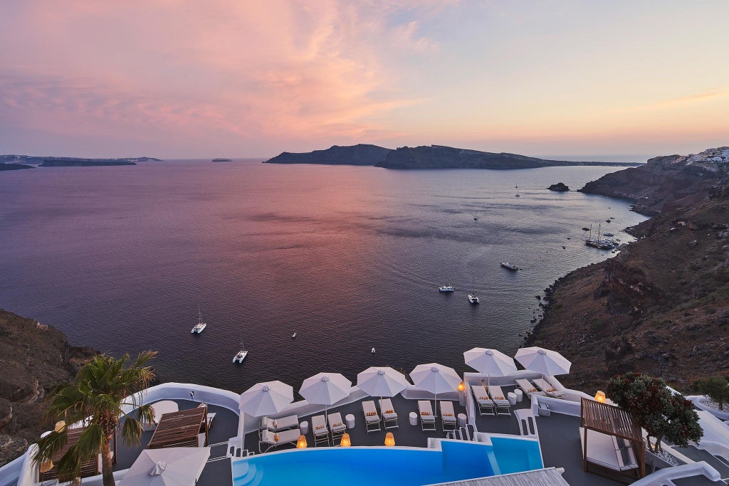 Luxurious white-washed suites of a Santorini hotel cascading down volcanic cliffs, overlooking the sparkling blue Aegean Sea at sunset