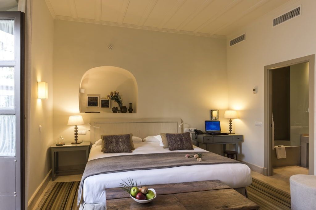 Elegant Spanish colonial-style hotel room with ornate wooden furniture, arched windows, and soft neutral tones overlooking historic Córdoba cityscape