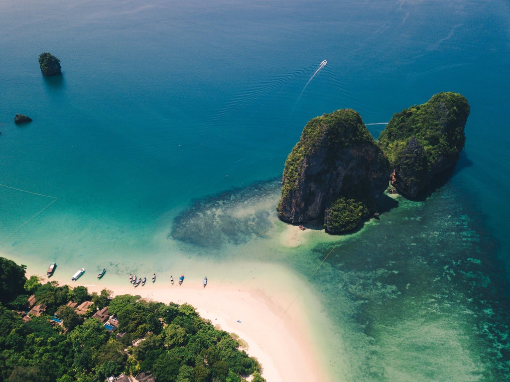 Steep limestone cliffs rise from turquoise waters, with traditional Thai longtail boats anchored near a pristine white sand beach in Krabi