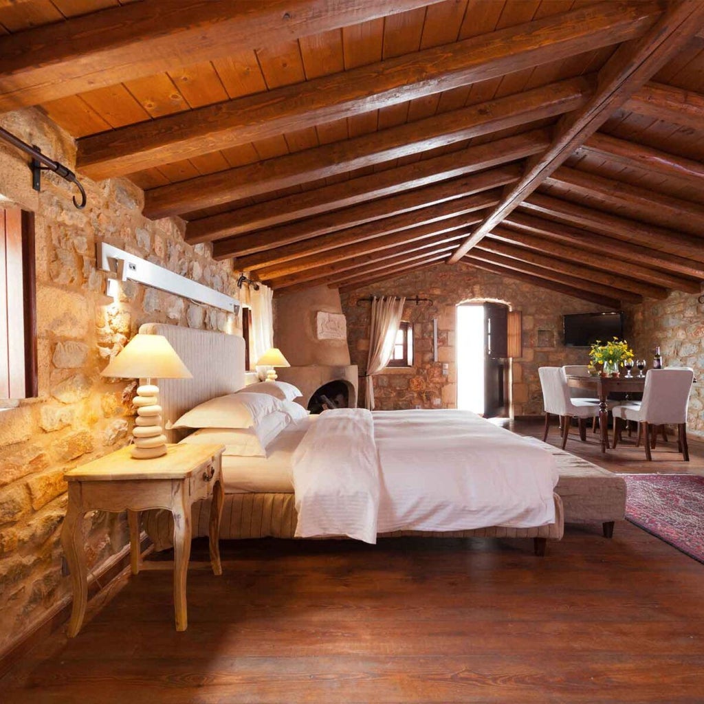 Elegant loft suite with stone walls, wooden beams, and plush white bedding overlooking a scenic coastal landscape in a traditional Greek hotel