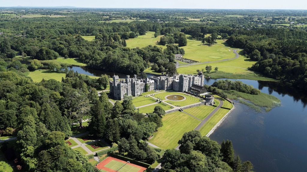 Medieval stone castle with ivy-covered walls, turrets and towers reflecting in a tranquil lake, set amid manicured gardens on a foggy Irish morning