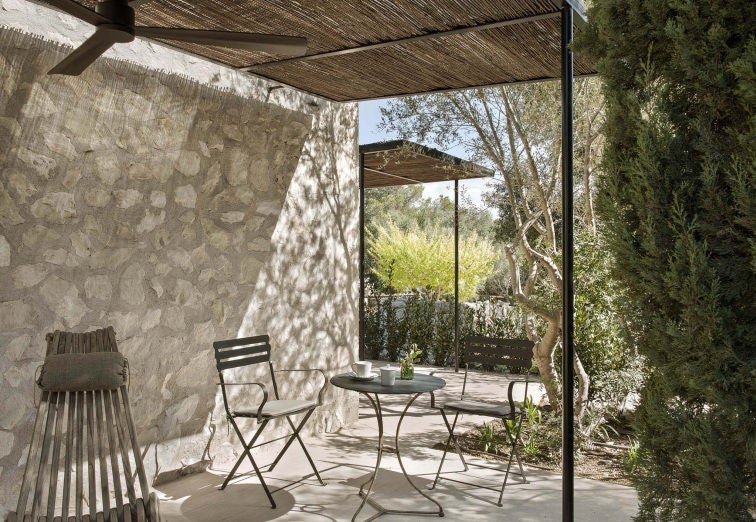 Serene white hotel room with king bed, rustic wooden beams, floor-to-ceiling windows overlooking Mediterranean garden and terrace.