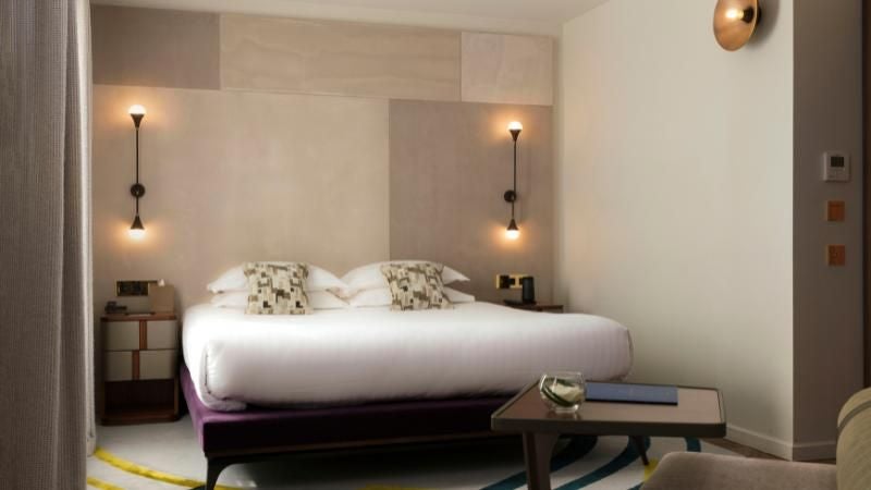 Elegant Parisian hotel room with plush bedding, soft neutral tones, designer accent chair, and large windows overlooking a chic urban landscape