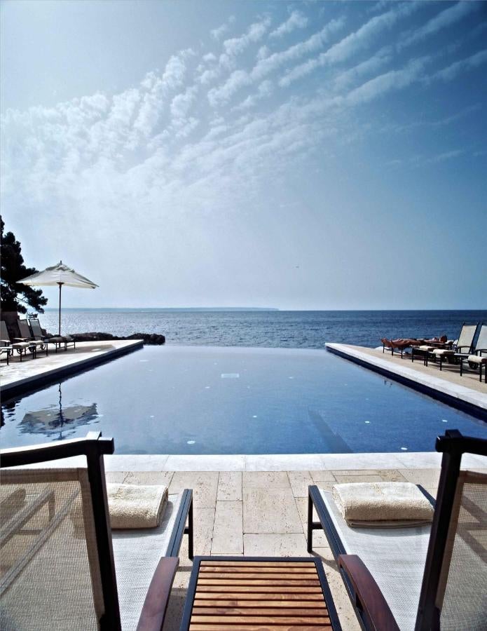 Elegant beachfront hotel with stone archways and terraces overlooking Mediterranean Sea at sunset, palm trees frame classic facade