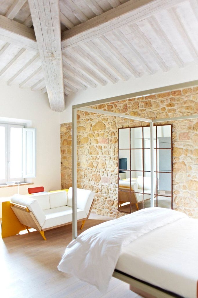 Elegant Tuscan hotel room with minimalist design, soft neutral tones, plush white bedding, rustic stone wall, and large windows overlooking scenic landscape