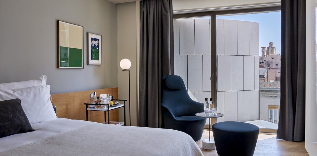 Spacious modern hotel room with minimalist design, sleek wooden furnishings, floor-to-ceiling windows overlooking Barcelona's urban landscape, elegant neutral color palette