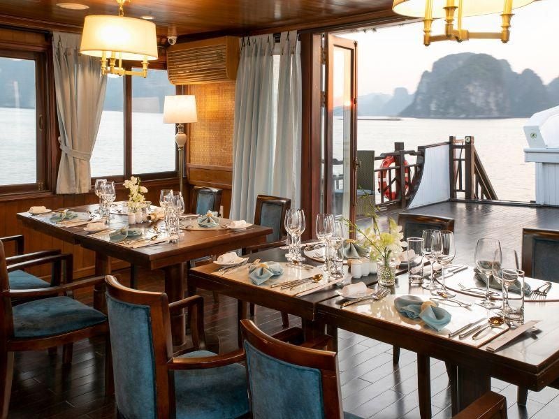 Elegant wooden-paneled cabin with traditional Vietnamese design, plush queen bed, rich mahogany furnishings, and panoramic window overlooking Halong Bay's emerald waters