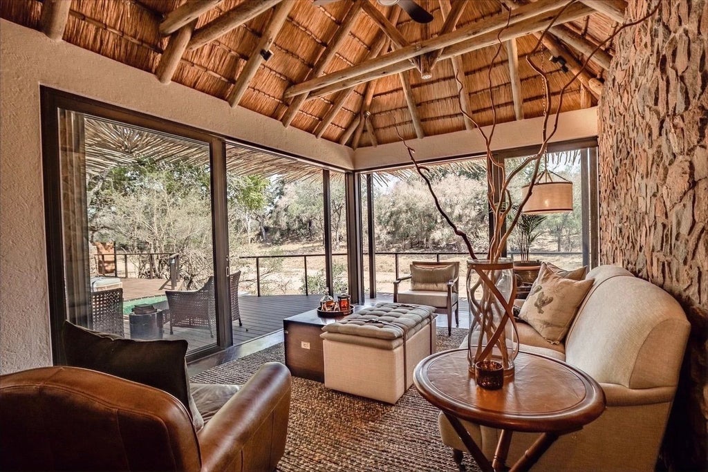 Elegant safari lodge with thatched roof and wooden deck overlooking a serene waterhole, surrounded by African bushveld at sunset