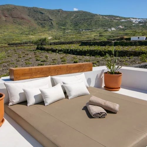 Elegant Greek villa bedroom with rustic stone walls, plush white bedding, arched window overlooking scenic vineyard landscape at dusk