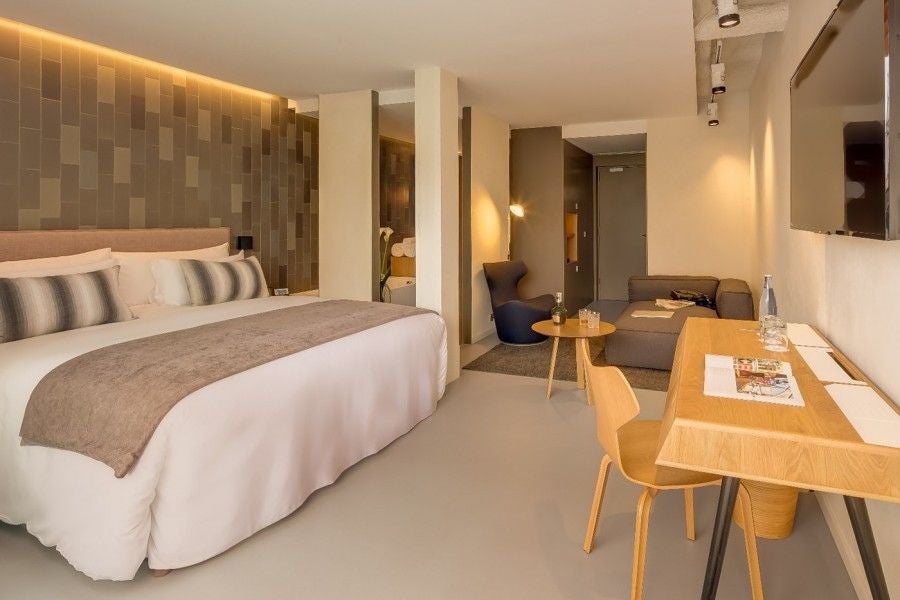 Elegant modern hotel room with minimalist design, crisp white linens, sleek furniture, and large windows overlooking urban Barcelona landscape.