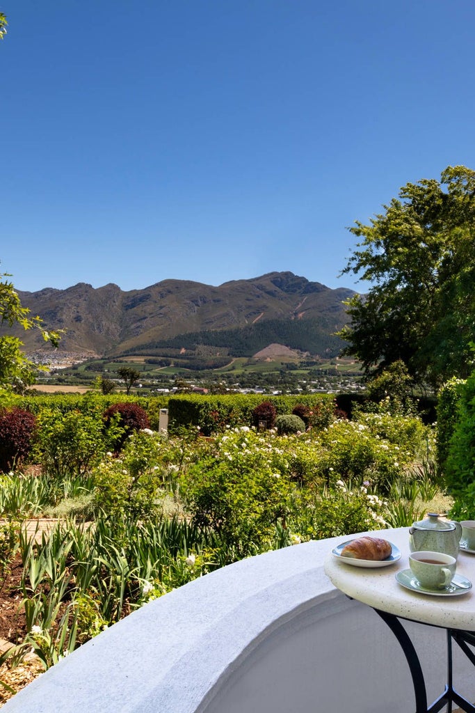 Luxurious South African wine country suite with plush white bed, elegant wooden furnishings, and expansive vineyard views through floor-to-ceiling windows